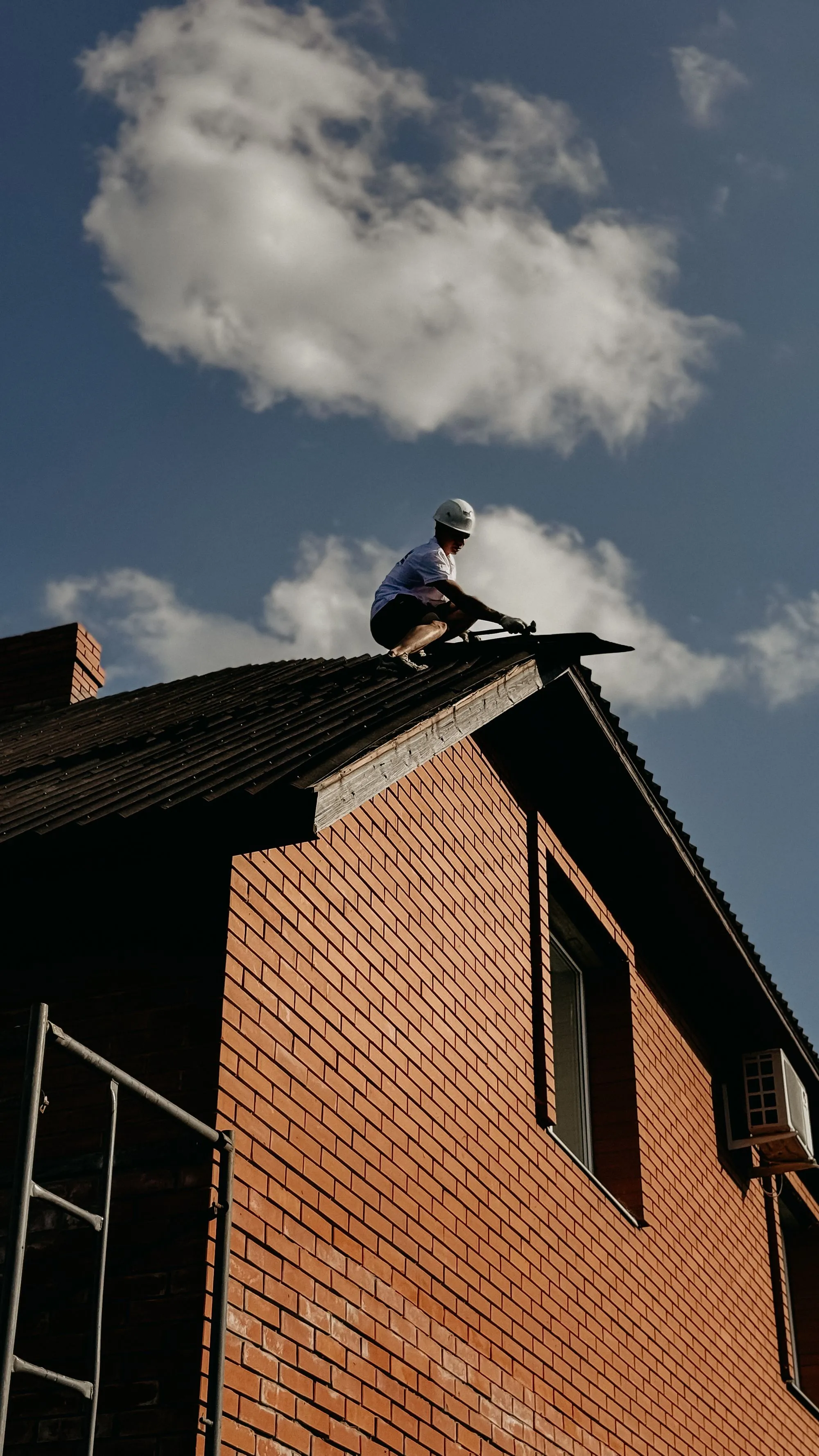 A roofer working along the ridge of a steep residential roof against open sky